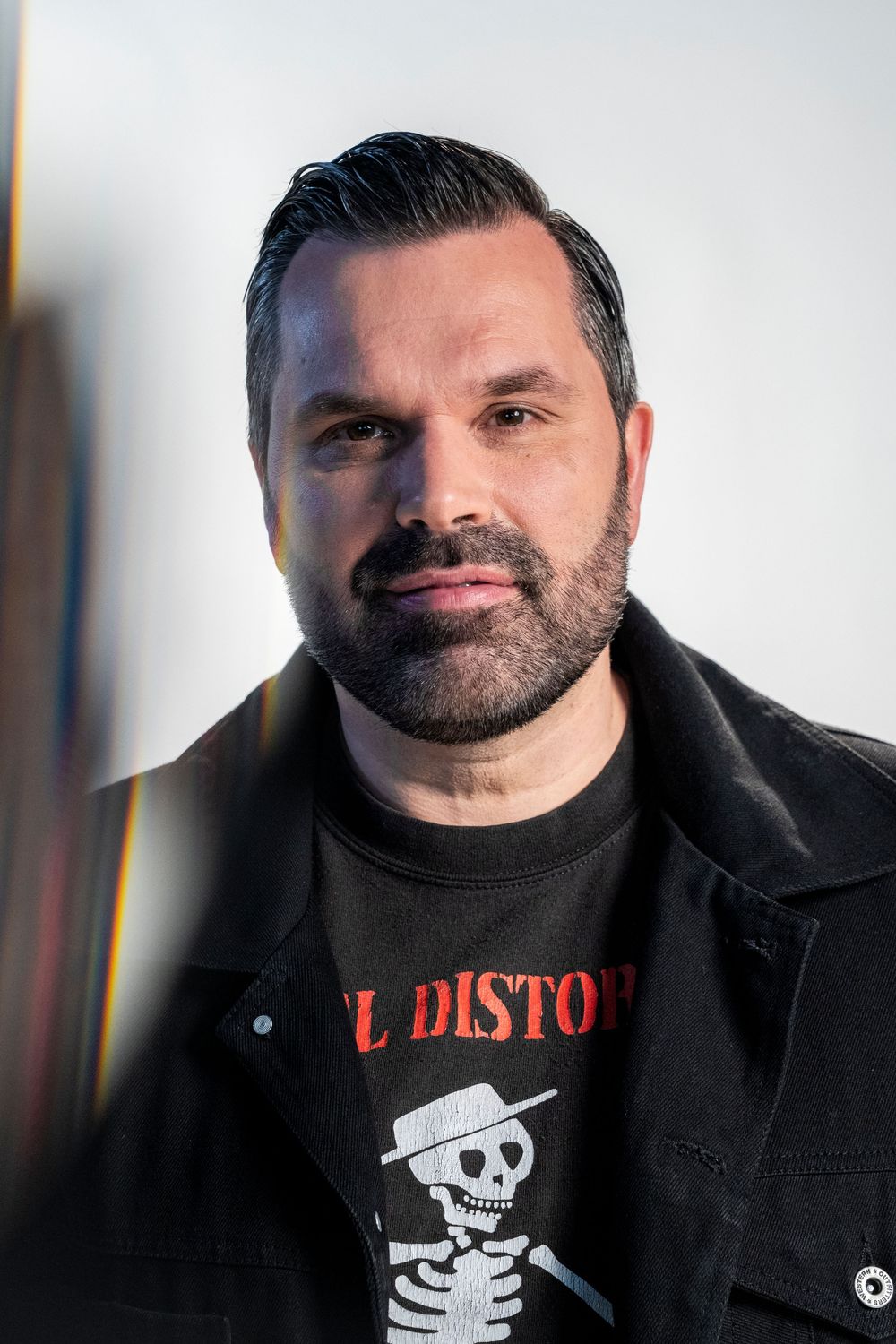 A bearded man in a black jacket and skull graphic tee poses confidently against a bright white background.