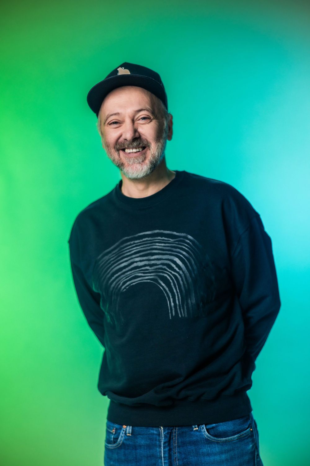 Smiling middle-aged man with beard wearing dark sweatshirt and cap poses against vibrant green gradient background.