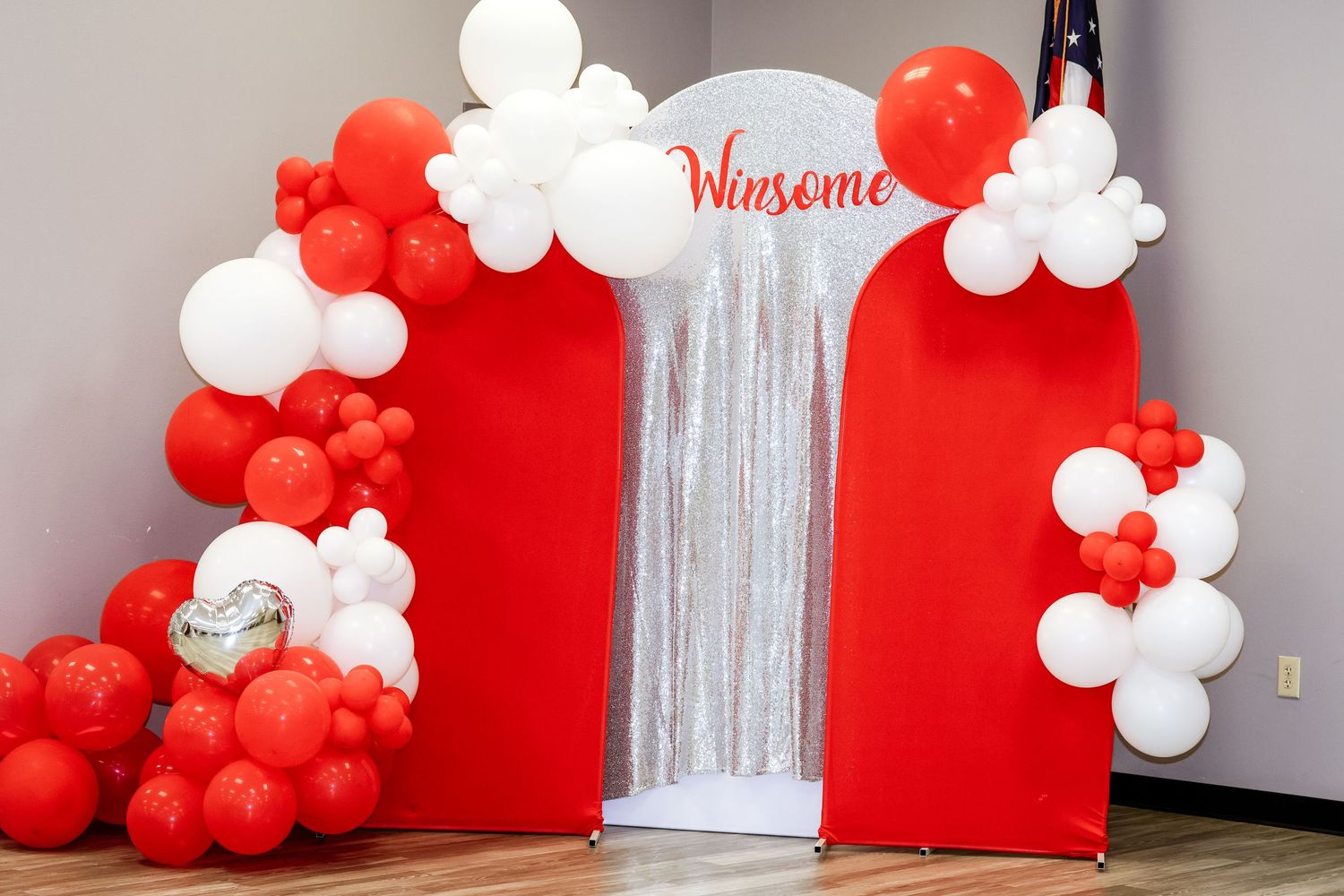 Red and white balloon arch with silver tinsel backdrop displaying Winsome sign at a festive event.