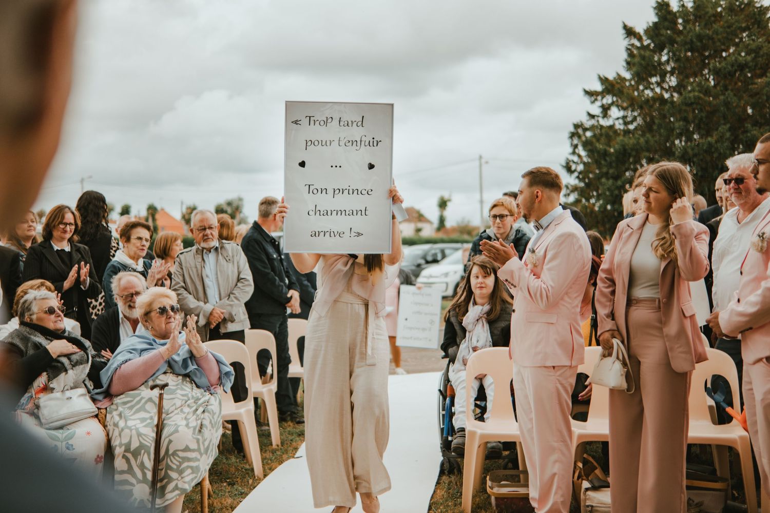 Outdoor wedding ceremony with guests seated as a person holds a French sign, while a couple in pink suits stands nearby.