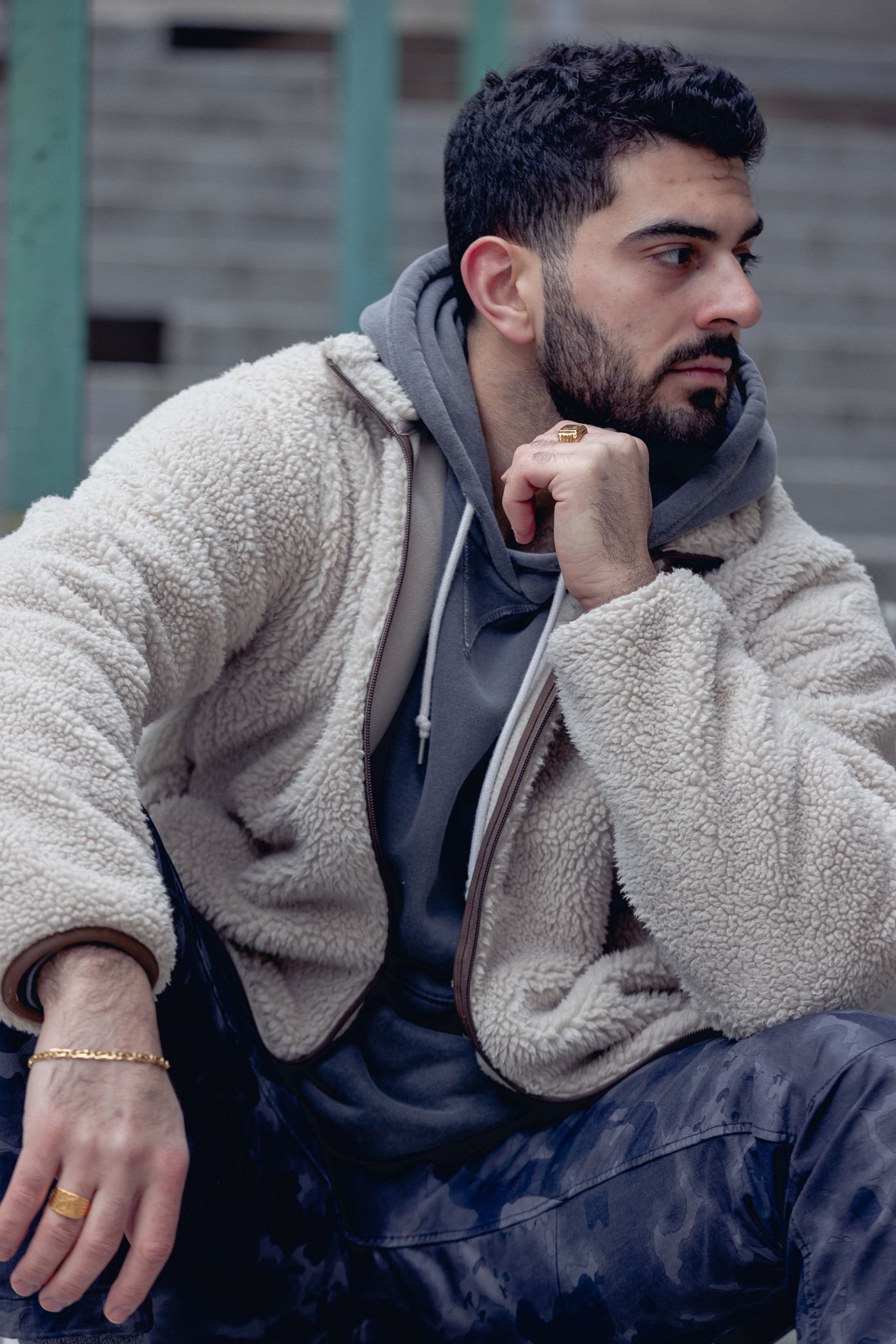 A bearded man in a sherpa jacket and gray hoodie sits thoughtfully outdoors, wearing gold rings and a bracelet.