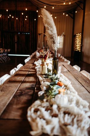 Rustic barn wedding reception table with pampas grass centerpieces, macrame runner, and string lights overhead.