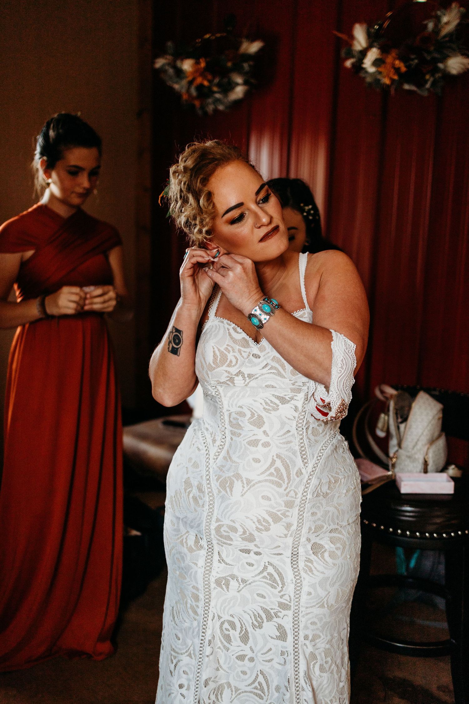 Bride in white lace gown putting on earrings with bridesmaid in rust dress in background.