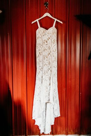 White lace wedding dress hanging on a wooden hanger against a rustic red barn wall.