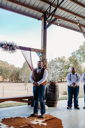 Groom in brown vest and jeans stands at rustic barn wedding altar with barrel decor and groomsmen nearby.