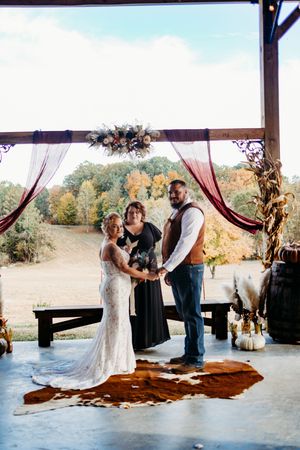 Bride and groom exchange vows at a rustic barn wedding ceremony with burgundy drapes and fall foliage backdrop.