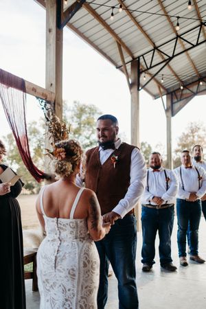 Bride and groom hold hands during outdoor barn wedding ceremony with groomsmen in suspenders behind them.