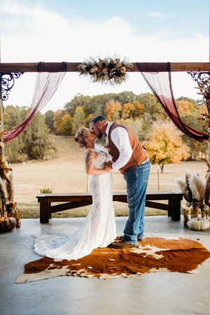 Bride and groom sharing first kiss under decorated arch with burgundy draping and fall florals on cowhide rug.