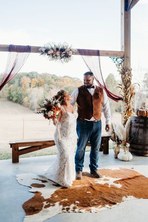 Bride and groom celebrate after ceremony under floral arch with cowhide rug and autumn hillside backdrop.