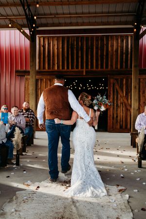 Bride and groom share first kiss at rustic barn wedding ceremony with string lights and seated guests.