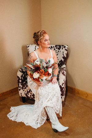 Bride in white lace gown and cowboy boots holding fall bouquet seated in cowhide chair.
