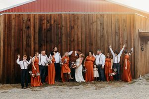 Wedding party celebrates in front of a red barn, bridesmaids in rust dresses and groomsmen in suspenders.