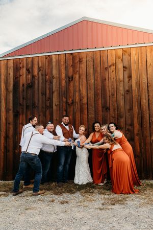 Wedding party in rust orange dresses and suspenders posing and laughing against a wooden barn wall.