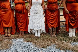 Bride in lace gown with bridesmaids in burnt orange dresses and cowboy boots at rustic wedding.