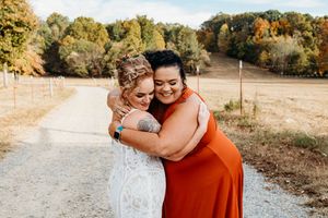 Bride in lace gown and bridesmaid in rust orange dress share a warm embrace on a country dirt road.