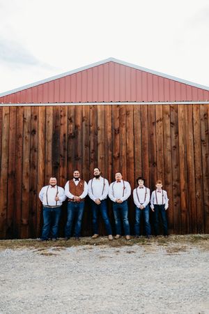 Groomsmen in white shirts, suspenders, and jeans posing in front of a rustic wooden barn with red roof.