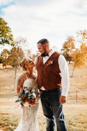 Bride and groom share a tender moment outdoors during fall wedding, she holds a rustic bouquet as autumn trees glow behind them.