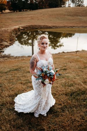 Bride in lace wedding gown holding a rust and blue bouquet, standing by a pond on autumn grass.