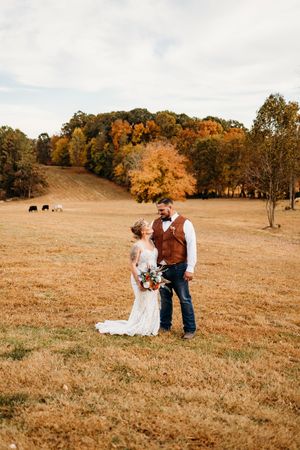 Bride and groom share a romantic moment in an autumn field with fall foliage and cattle in the background.