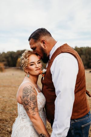 Groom in brown vest kissing bride on forehead in golden field, bride wearing lace dress with rose tattoo sleeve.