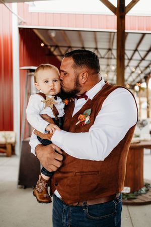 A groom in a brown vest kisses a baby boy at a rustic wedding venue.