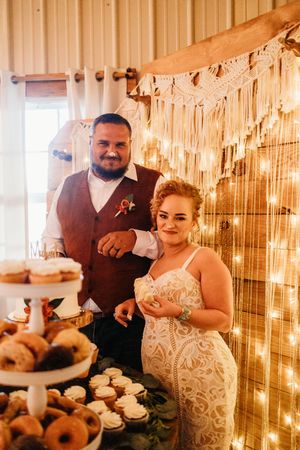 Bride and groom smiling at wedding cake table with donuts, string lights, and macrame backdrop in rustic barn venue.