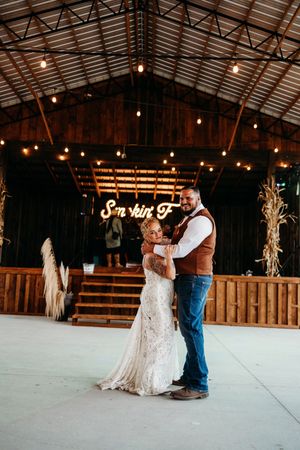 Bride and groom share first dance in rustic barn venue with string lights and neon sign backdrop.