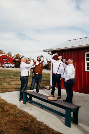 Groomsmen in white shirts and suspenders taking shots outside a rustic red barn at a country wedding.