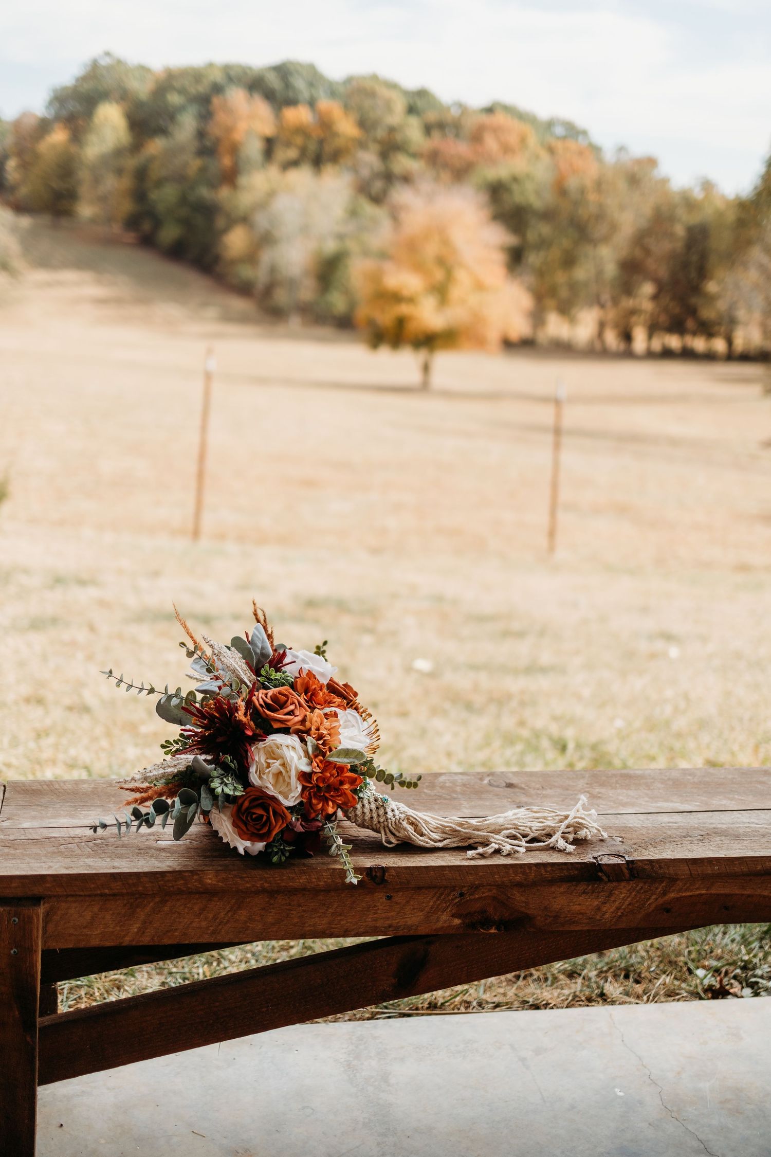 Autumn wedding bouquet with burnt orange and ivory roses on a rustic wooden bench, set against a golden fall landscape.
