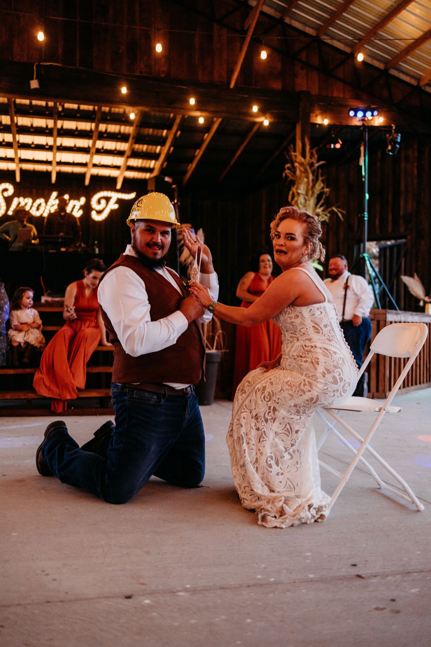 Groom kneels on dance floor as bride places hat on his head during fun wedding reception garter tradition.