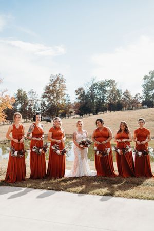 Bride in lace gown stands with bridesmaids in rust orange dresses holding bouquets at an outdoor fall wedding.