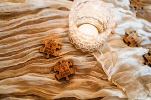 Rustic wooden puzzle piece wedding favors with engraved text beside a glowing candle on cream cheesecloth table runner.