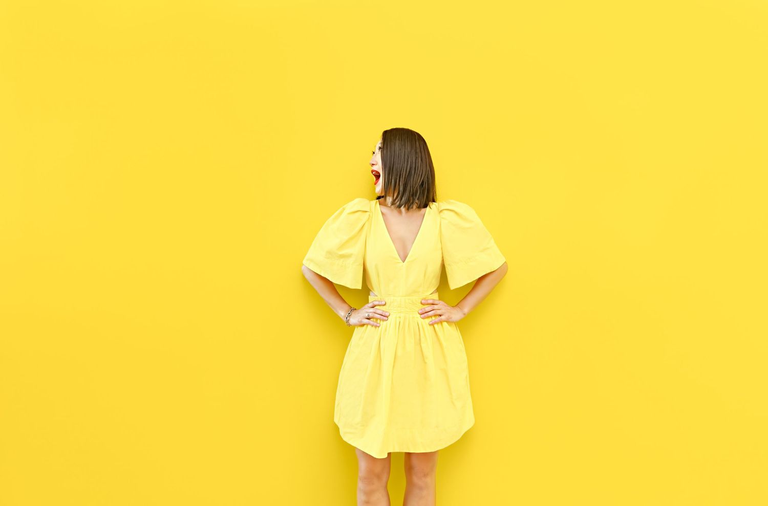 Woman in yellow puff-sleeve mini dress poses against bright yellow wall, hands on hips, looking sideways.