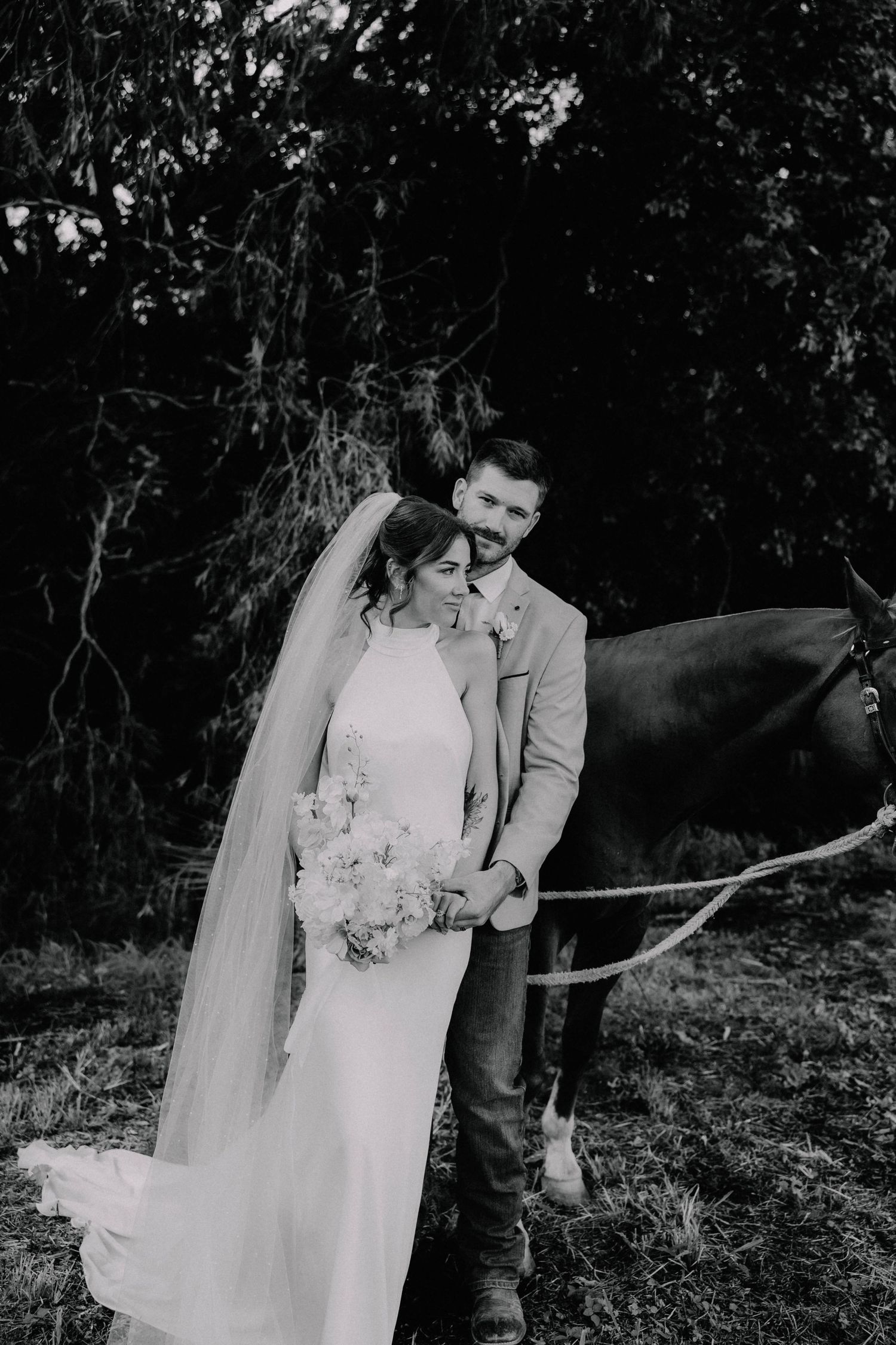 Bride in white backless gown and groom in tan blazer embrace near a sunlit pond surrounded by lush green trees.