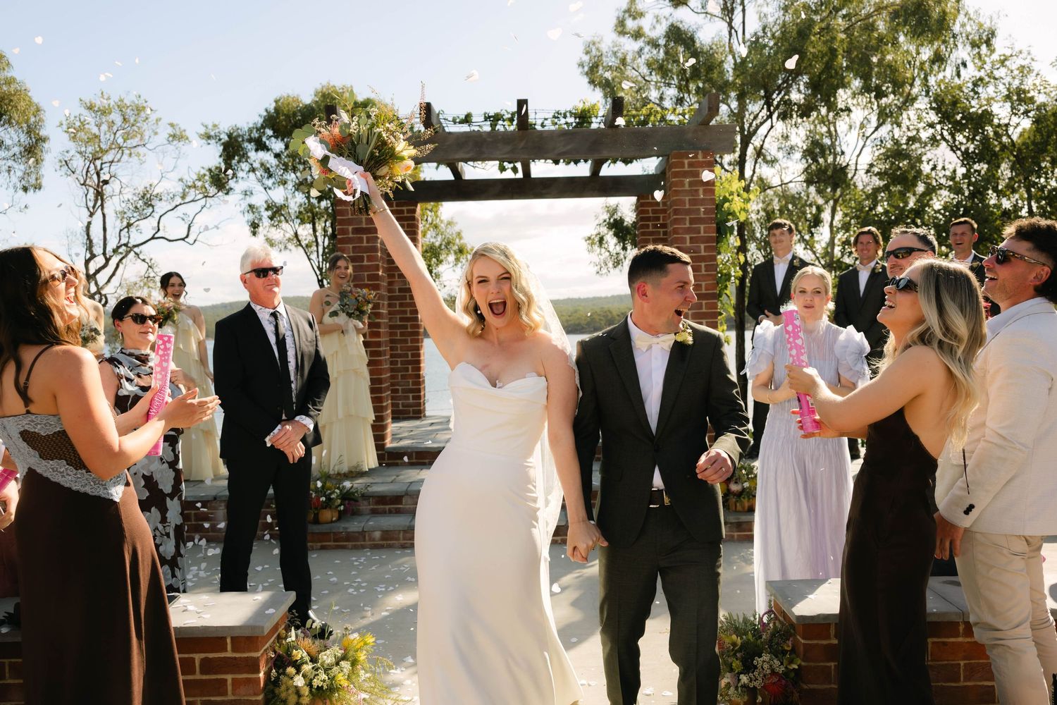 Bride in white backless gown and groom in tan blazer embrace near a sunlit pond surrounded by lush green trees.