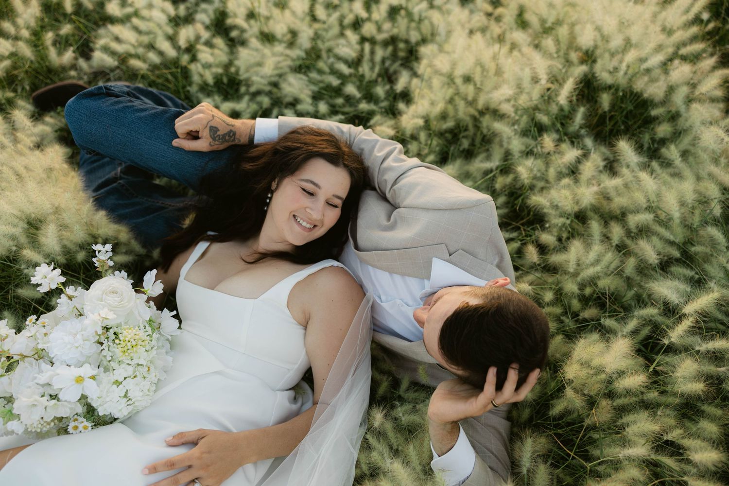 A bride and groom lying in a lush green field, smiling and holding a white floral bouquet on their wedding day.