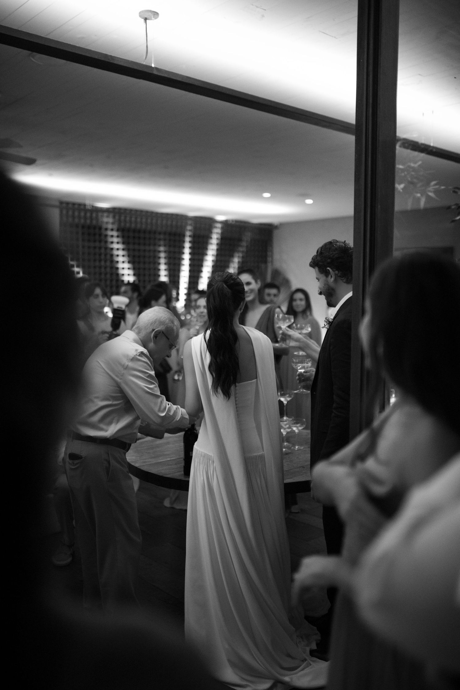 Black and white photo of a bride in a white gown at a wedding reception surrounded by guests.