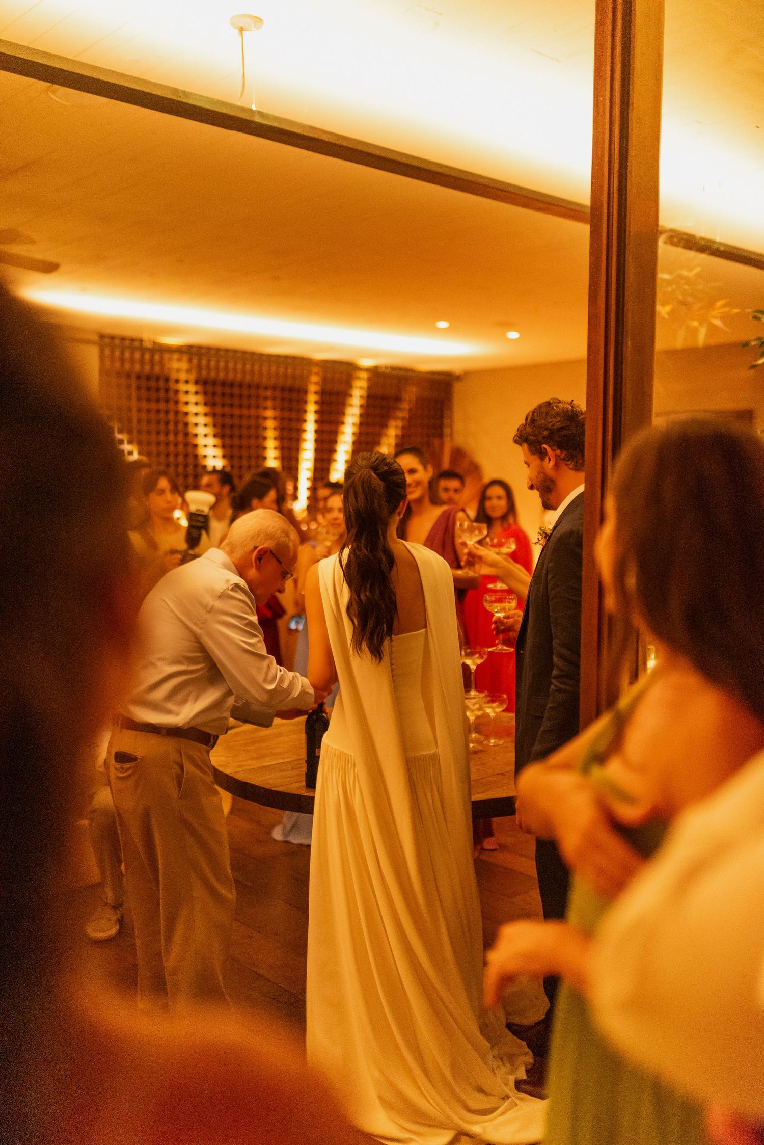 Bride in elegant white gown at a warmly lit wedding reception with guests in formal attire.