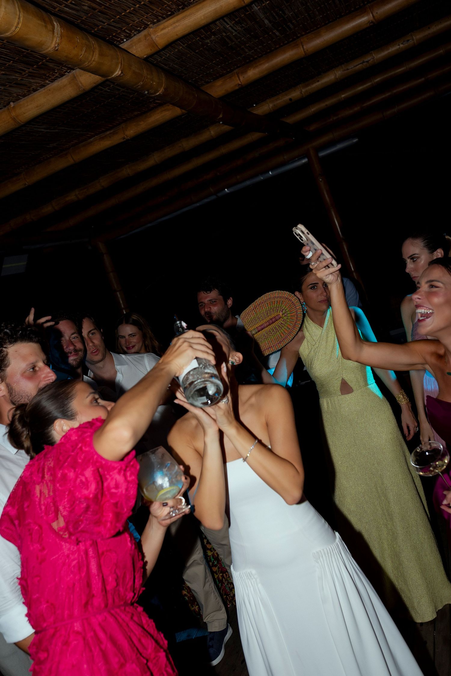 Bride in white dress drinks from large cocktail shaker at lively outdoor wedding reception party with guests.
