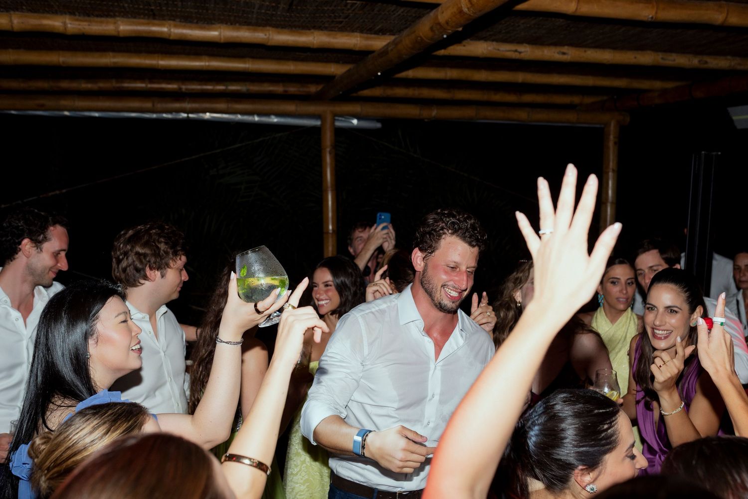 Guests dancing and raising glasses at a lively outdoor party under a thatched roof at night.