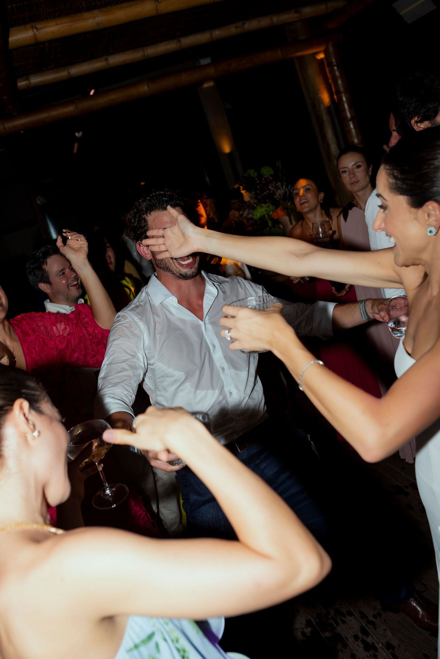 Guests laughing and dancing at a lively wedding reception party, with a woman playfully covering a man's eyes.