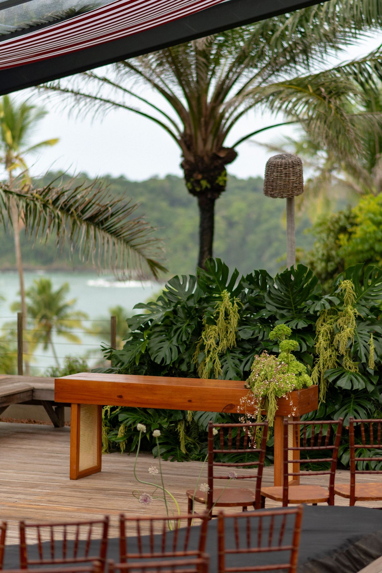 Tropical outdoor wedding venue with wooden table, monstera plants, palm trees, and ocean view on a deck.