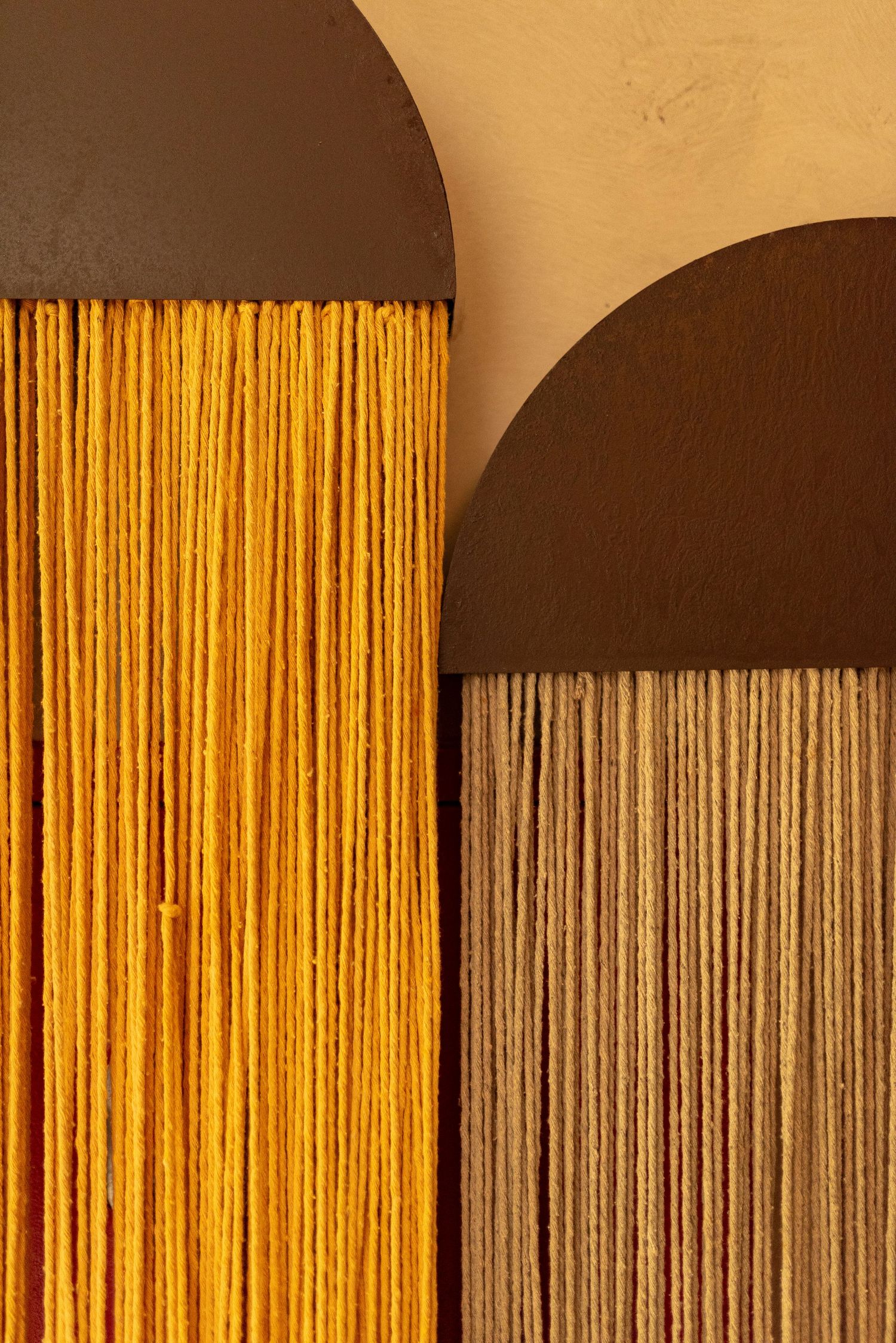 Two decorative wall hangings with dark brown arched tops and hanging orange and tan fiber threads on a beige background.