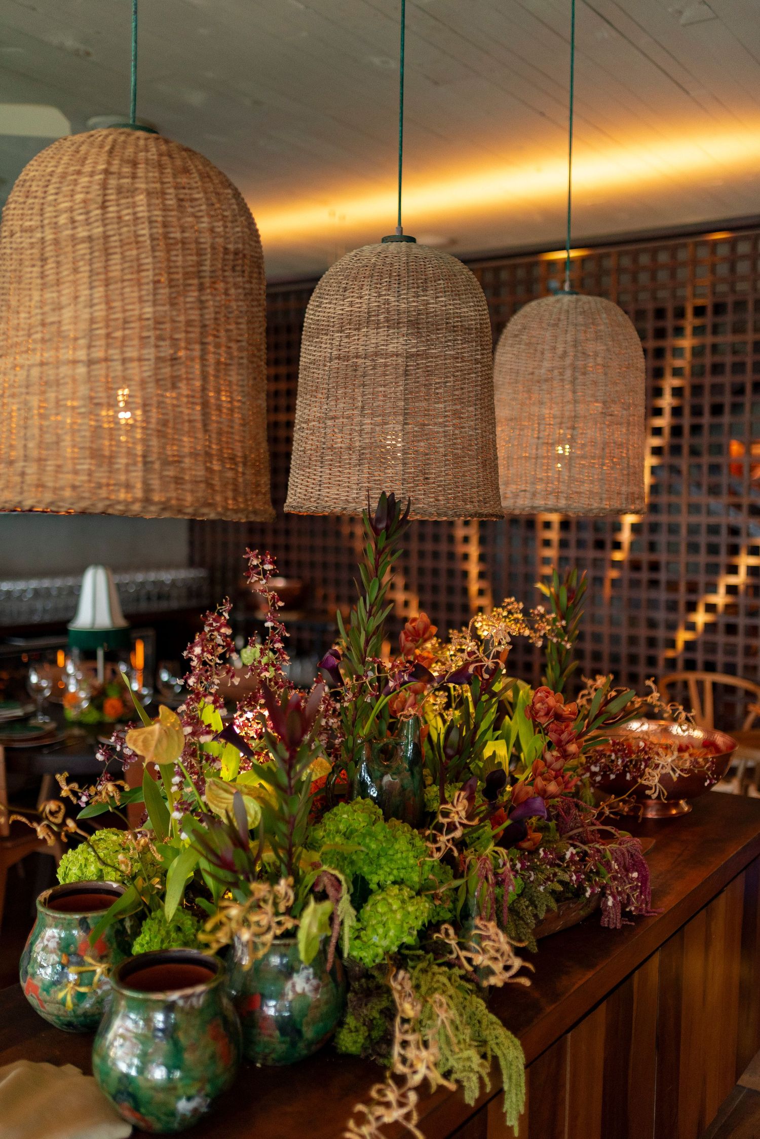 Wicker pendant lights hang over a wooden table with lush floral arrangements and colorful vases in a warmly lit restaurant.