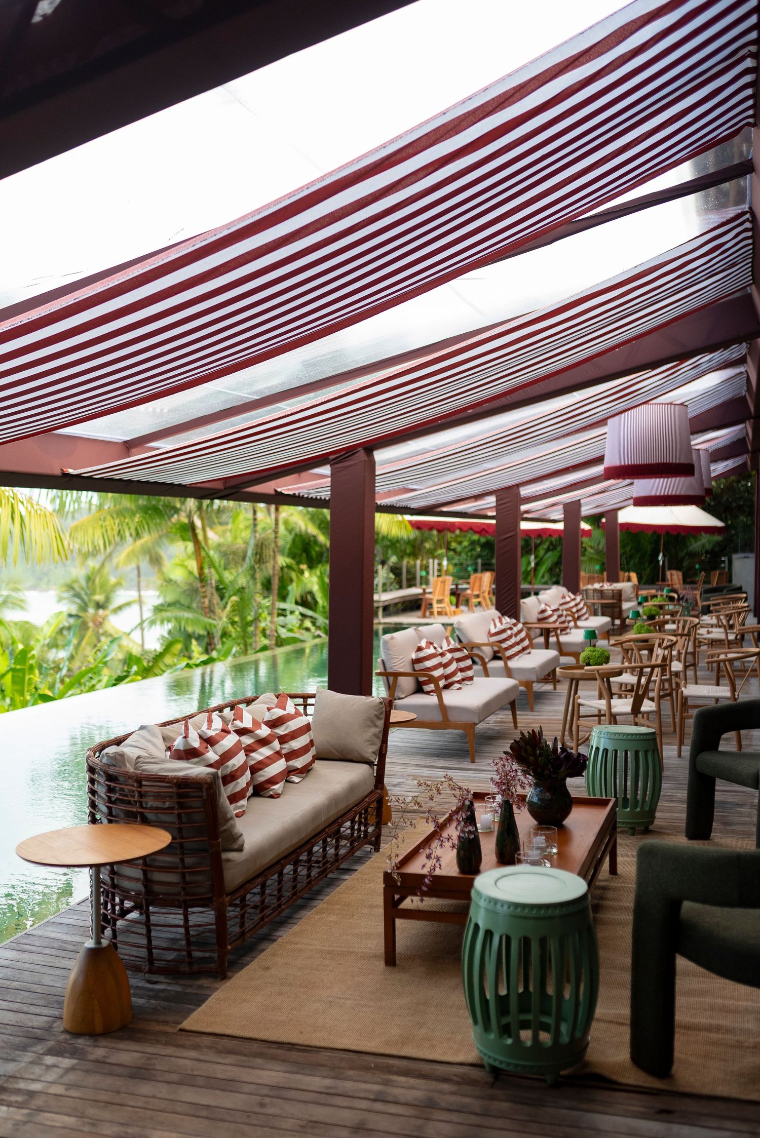 Tropical resort lounge with striped canopy, rattan furniture, green ceramic stools, and lush palm tree views beside a pool.