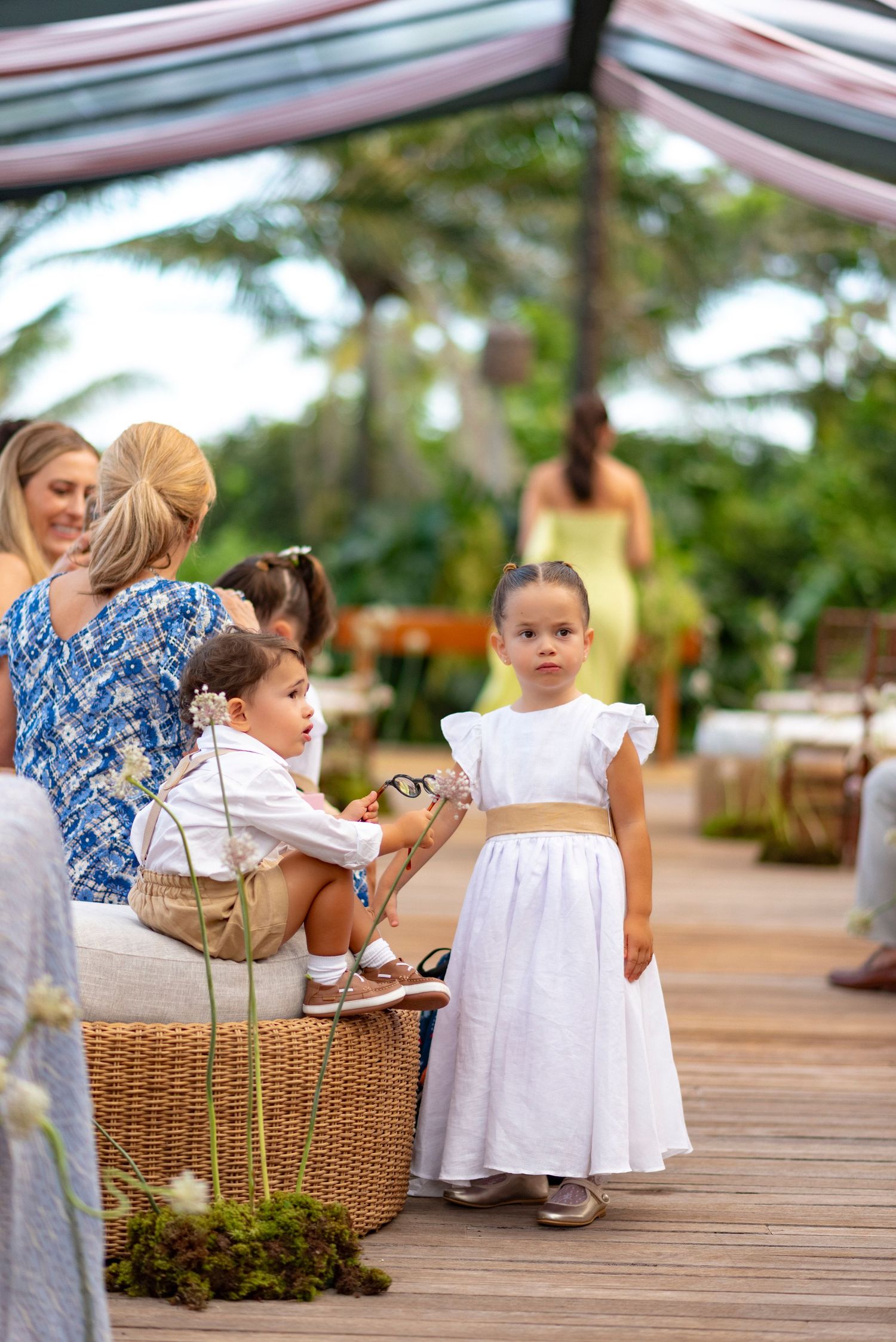A flower girl in a white dress stands on a wooden deck at an outdoor wedding ceremony surrounded by lush greenery.
