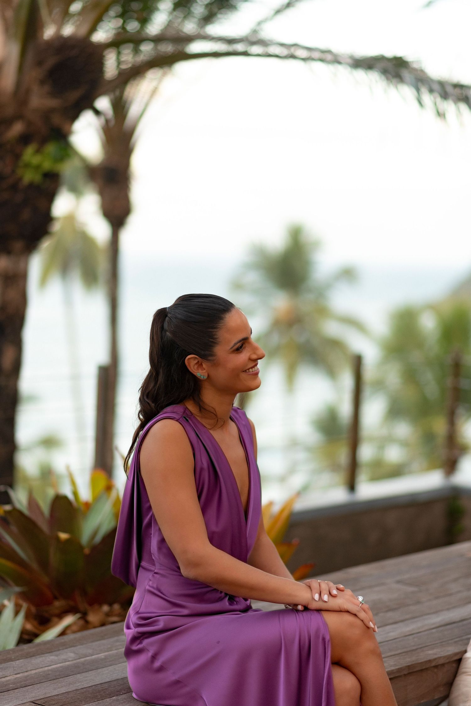 Woman in purple dress sitting outdoors near palm trees and ocean, smiling serenely at a tropical beach resort.