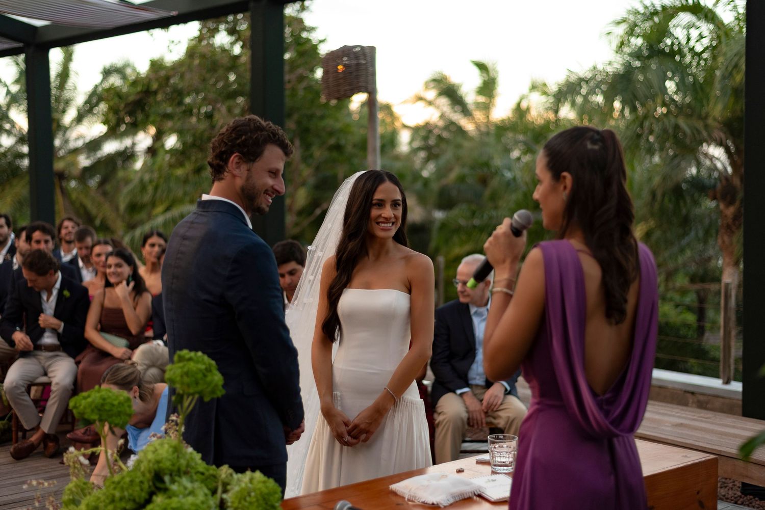 Bride in white gown and veil smiles at groom during outdoor wedding ceremony with guests seated behind them.