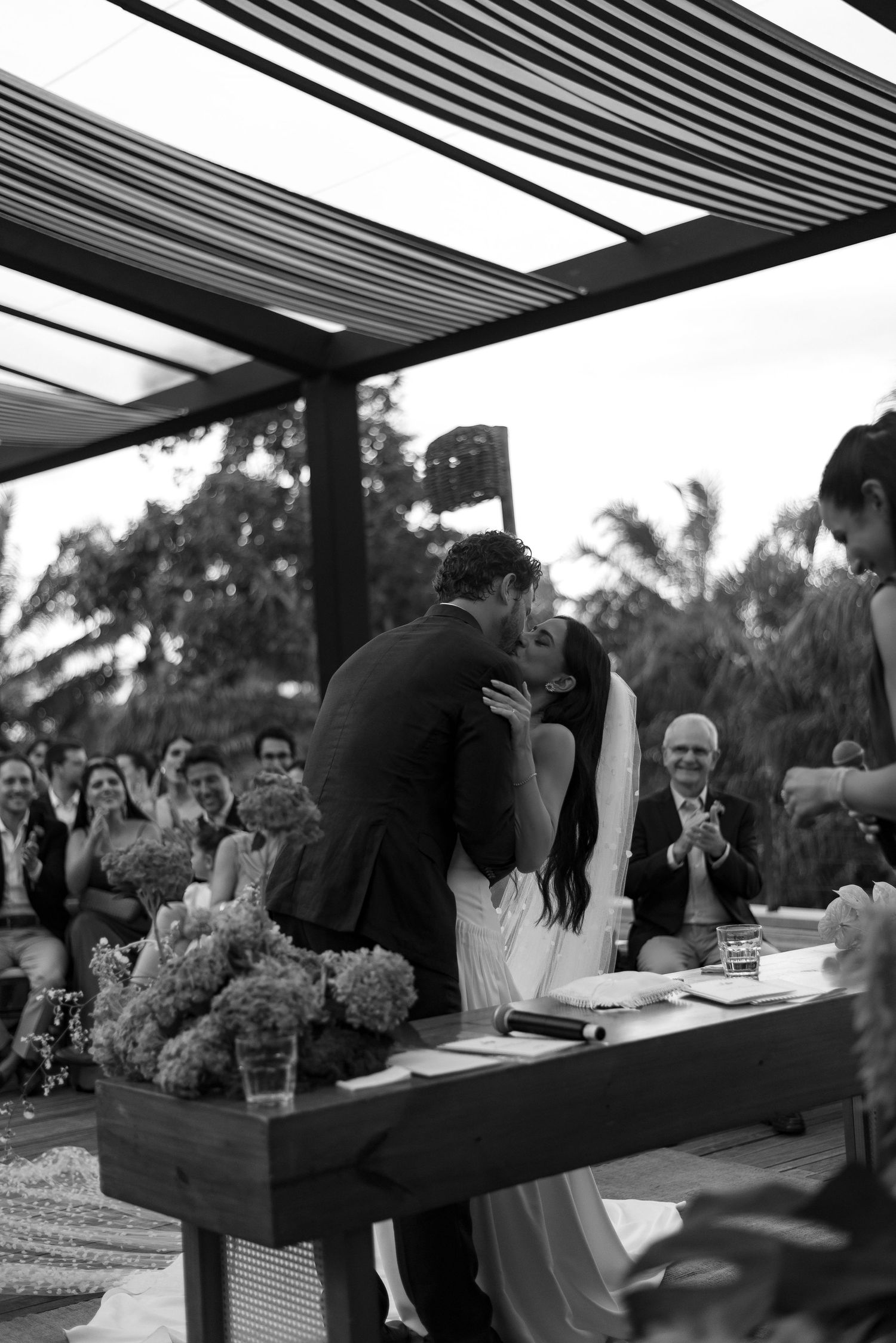 Black and white photo of a couple sharing their first kiss at an outdoor wedding ceremony with guests applauding.