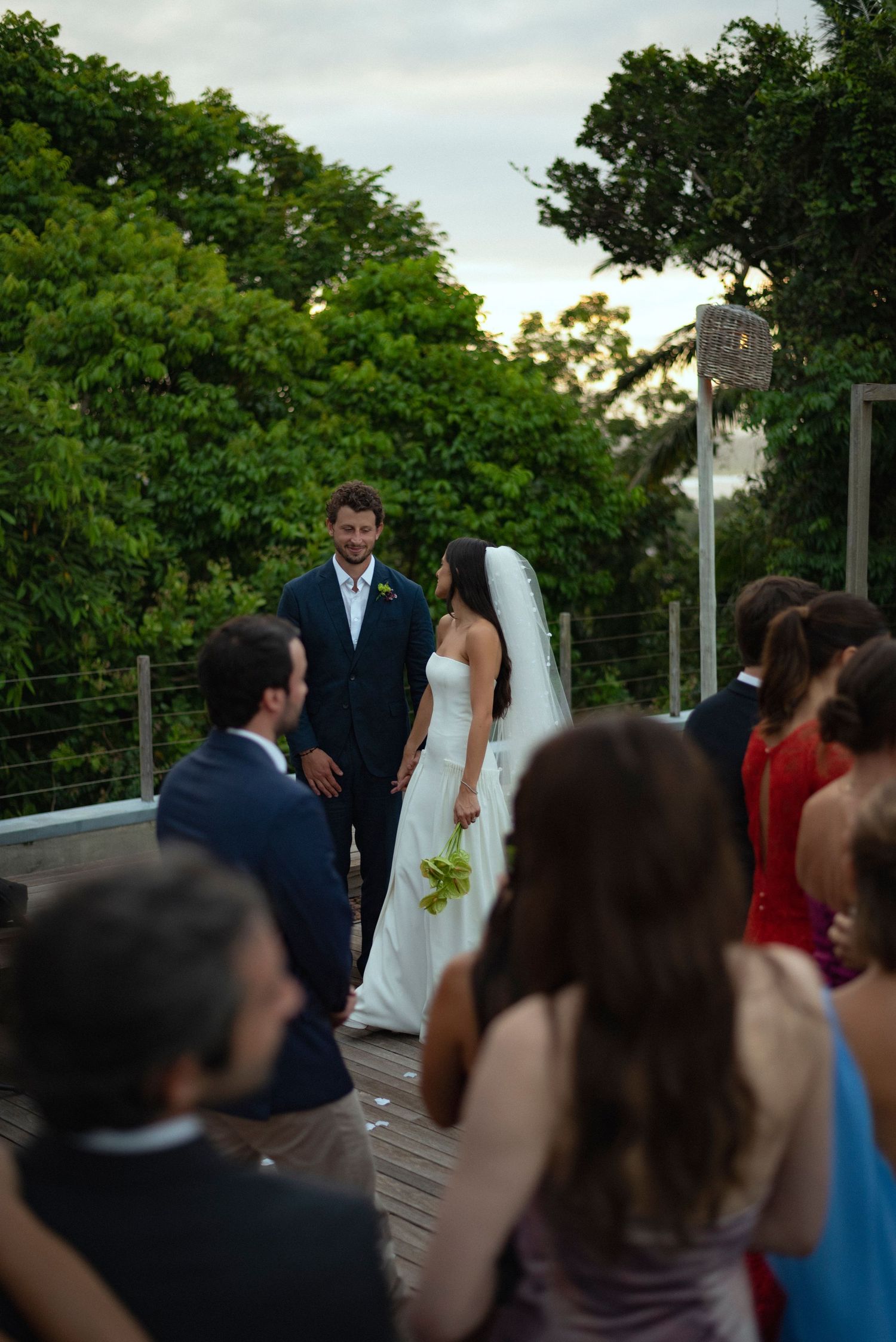 Outdoor wedding ceremony on a wooden deck with bride in white gown and groom in navy suit, surrounded by lush green trees.